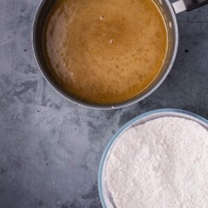 A mixing bowl of flour next to a pot containing a brown liquid batter, placed on a gray countertop.