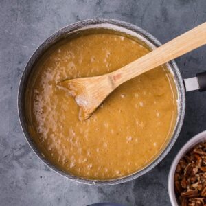 A saucepan filled with a thick, light brown mixture is being stirred with a wooden spatula; a bowl of pecans sits nearby.