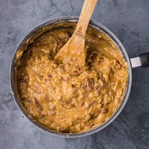 A saucepan filled with thick, creamy caramel pecan frosting being stirred with a wooden spatula on a gray surface.