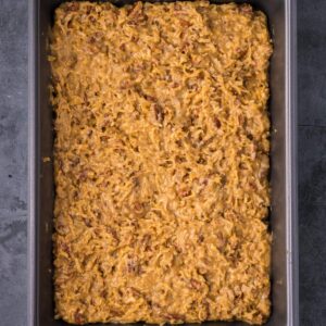 A rectangular baking dish filled with an uncooked mixture of shredded potatoes, cheese, and ground meat on a gray surface.