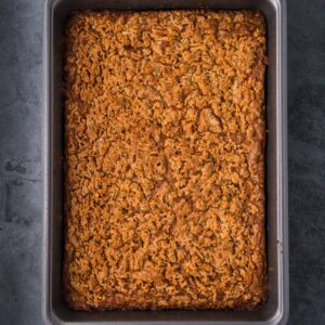 A rectangular baking pan containing a golden-brown baked oat and nut dessert, viewed from above on a dark surface.