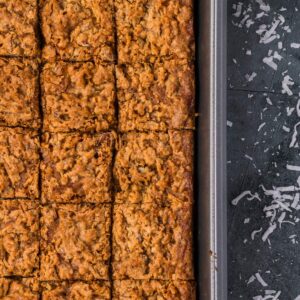 A baking pan of brownie bars cut into squares sits next to scattered shredded coconut on a dark surface.