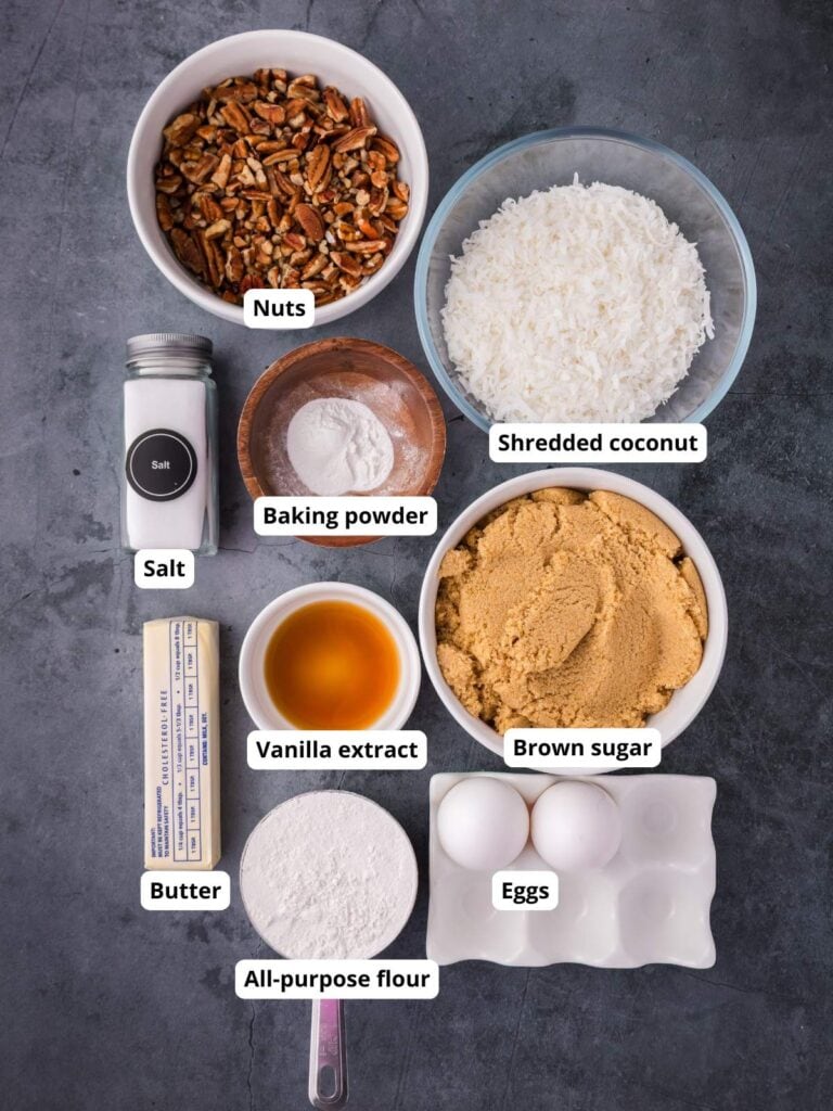 Overhead view of labeled baking ingredients in bowls and containers: nuts, shredded coconut, brown sugar, eggs, flour, butter, vanilla extract, salt, and baking powder.