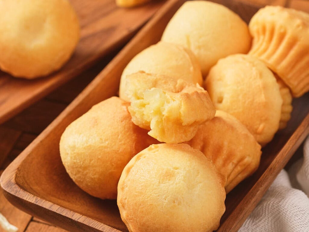 A wooden tray filled with round, golden-brown cheese breads, with one bread broken open to reveal its soft, airy inside.