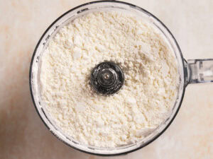 A food processor bowl filled with a white, powdery mixture, likely flour or blended dry ingredients, viewed from above.