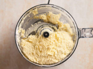 A top-down view of a food processor bowl filled with a crumbly, pale yellow mixture.