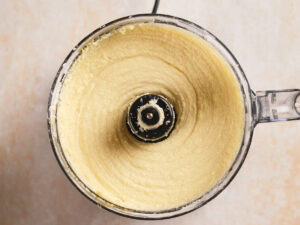 A food processor with smooth, creamy beige mixture inside, viewed from above on a light-colored surface.