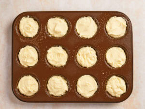 A muffin tin filled with twelve unbaked pastry shells containing a creamy batter, placed on a light-colored surface.