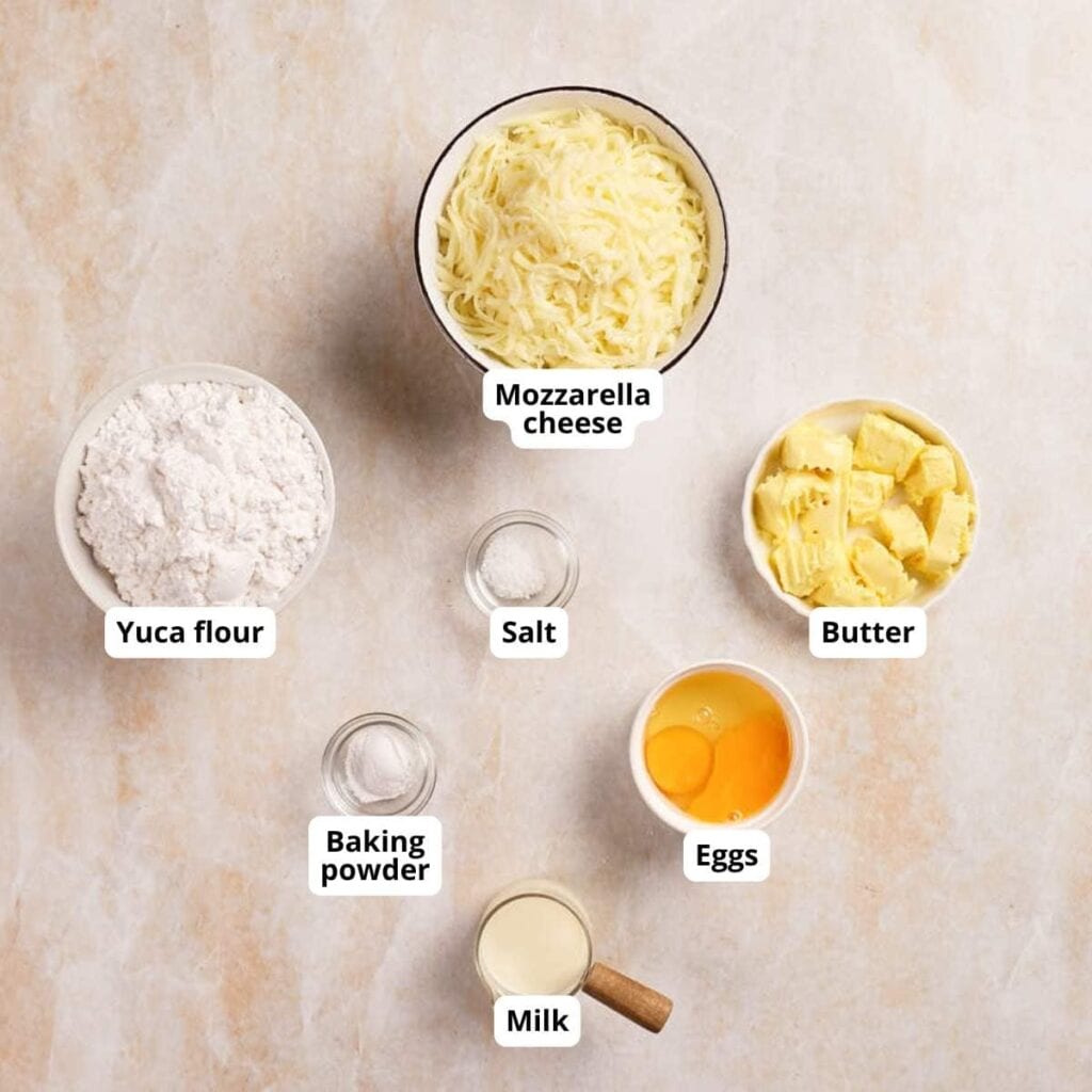 Six labeled ingredients on a light surface: yuca flour, mozzarella cheese, butter, eggs, salt, baking powder, and milk in separate bowls and containers.