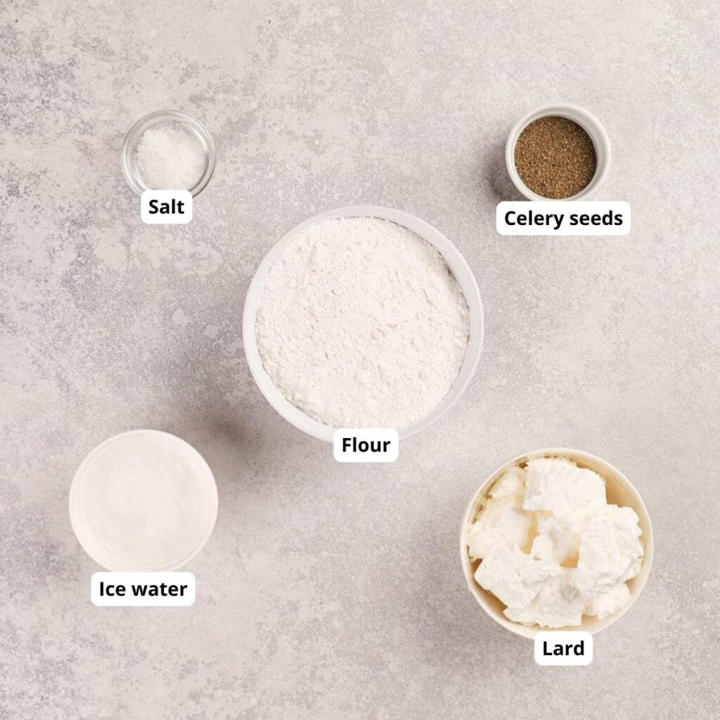 Five labeled bowls on a light surface containing flour, lard, ice water, salt, and celery seeds, arranged separately for a recipe ingredient display.