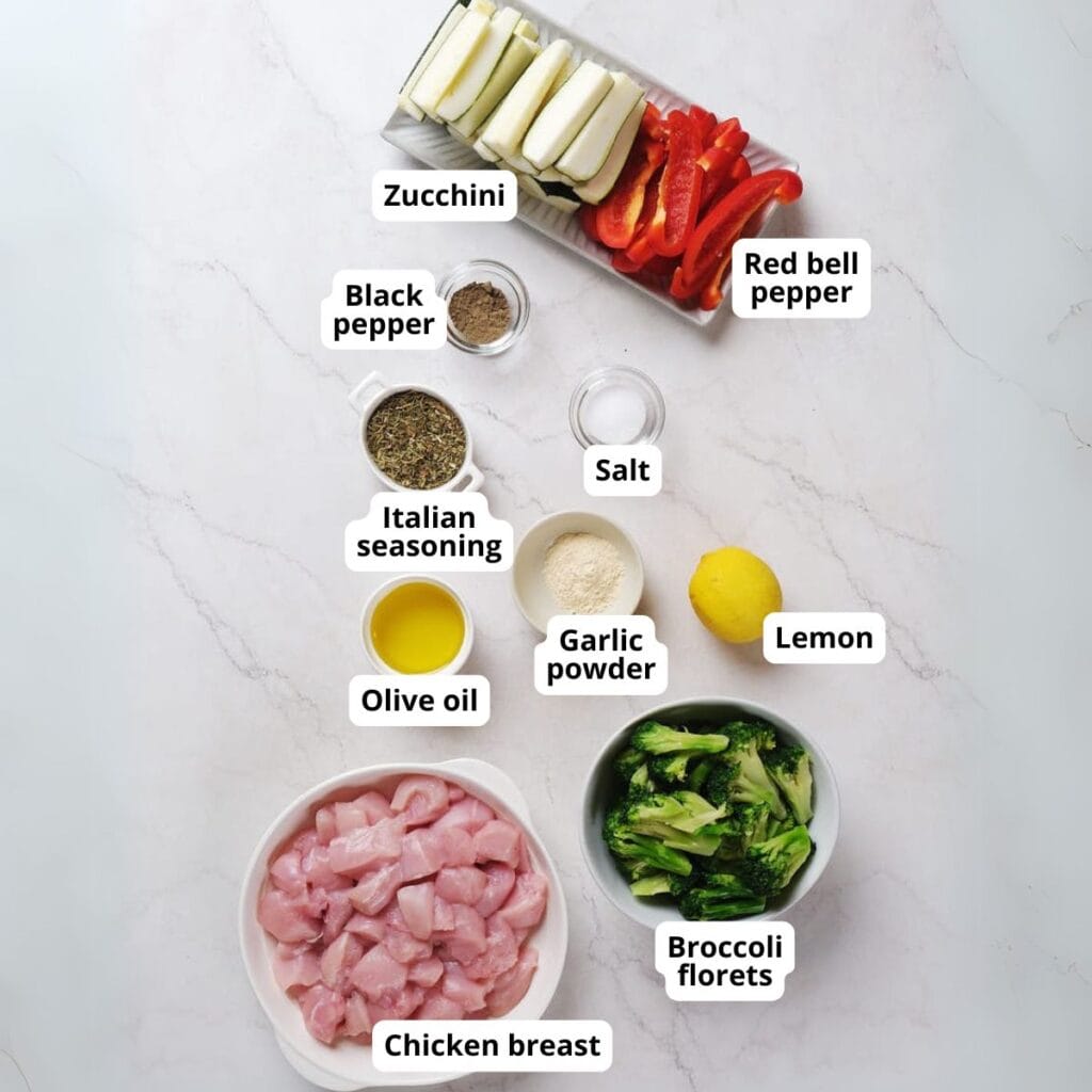 Chopped chicken breast, broccoli florets, zucchini, red bell pepper, olive oil, lemon, black pepper, Italian seasoning, salt, and garlic powder arranged on a white surface.