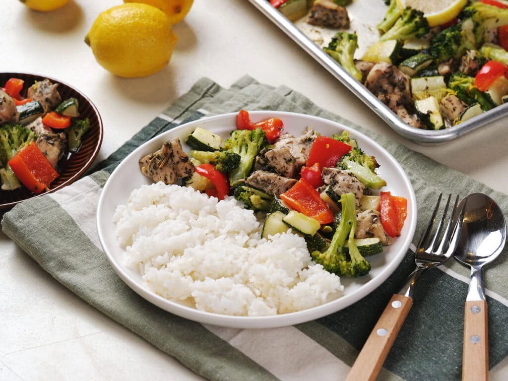 A plate with white rice and roasted chicken with broccoli, red bell pepper, and zucchini sits on a napkin, with a fork and spoon beside it.