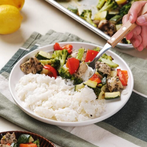 A bowl of white rice with roasted vegetables and chicken; a hand holds a fork with a bite of chicken and vegetables above the bowl.