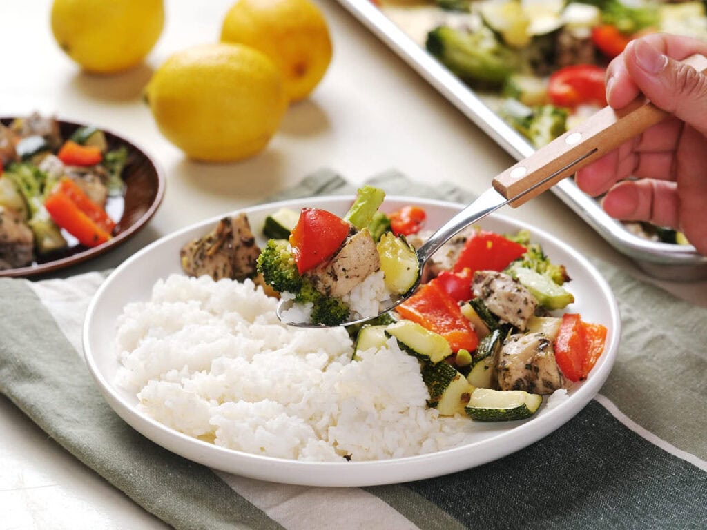 A hand spoons cooked vegetables and diced chicken onto a plate with white rice; lemons and a baking tray are in the background.