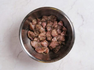 Raw chicken pieces coated in herbs and spices are mixed in a stainless steel bowl on a light marble surface.