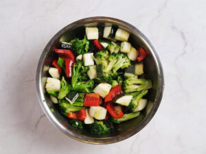 A stainless steel bowl filled with chopped broccoli, red bell peppers, and zucchini pieces on a light marble surface.