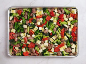 A baking sheet filled with diced chicken, broccoli, zucchini, and red bell pepper, all seasoned and ready to be cooked.