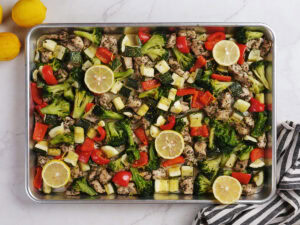 A sheet pan filled with chopped chicken, broccoli, zucchini, red bell pepper, and sliced lemons on a marble surface next to two lemons and a striped towel.