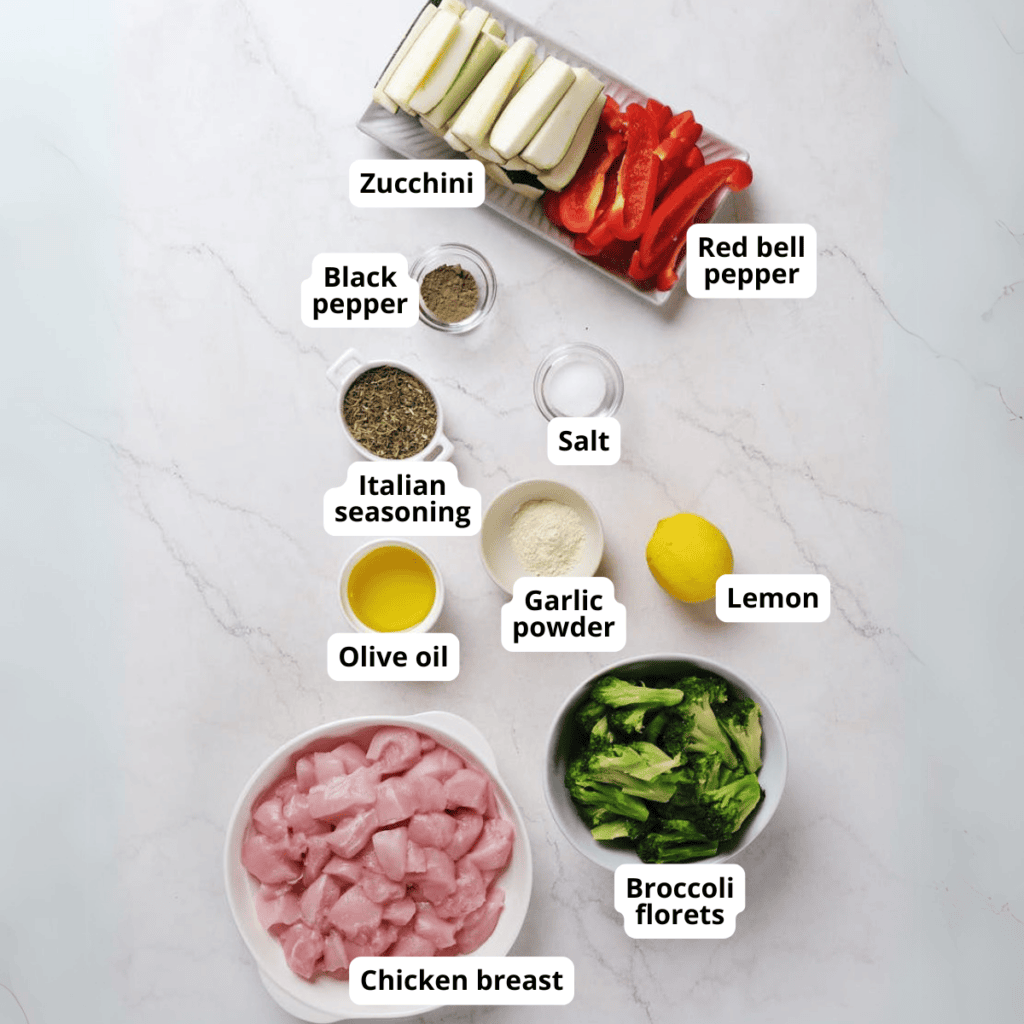 Chopped chicken breast, broccoli florets, zucchini, red bell pepper, olive oil, lemon, black pepper, Italian seasoning, salt, and garlic powder arranged on a white surface.