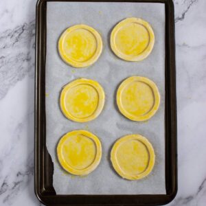 Six round, uncooked pastry circles brushed with egg wash are arranged on a parchment-lined baking sheet, ready for baking.