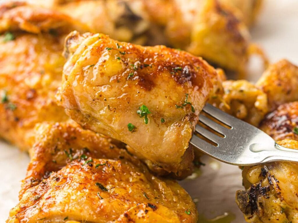 A close-up of a fork holding a piece of baked or roasted chicken thigh with a golden, crispy skin and sprinkled herbs.