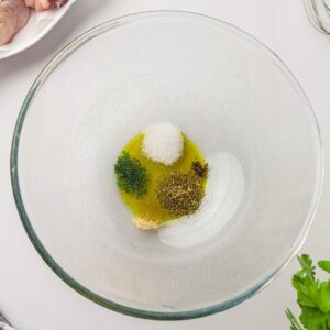 A glass bowl containing olive oil, salt, black pepper, dried herbs, and garlic powder, viewed from above on a white surface.