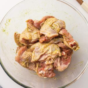 Raw chicken pieces coated with marinade in a clear glass mixing bowl, placed on a white surface.