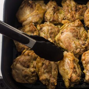 Pieces of cooked chicken are arranged in an air fryer basket, with a pair of metal tongs gripping one piece.