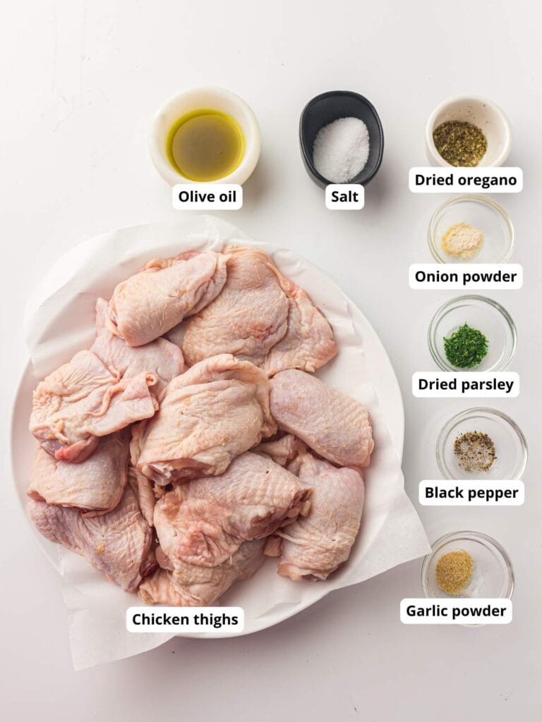 Raw chicken thighs on a plate surrounded by small bowls of olive oil, salt, dried oregano, onion powder, dried parsley, black pepper, and garlic powder.