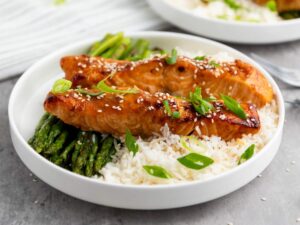 A plate with glazed salmon fillets topped with sesame seeds and green onions, served over white rice and asparagus.