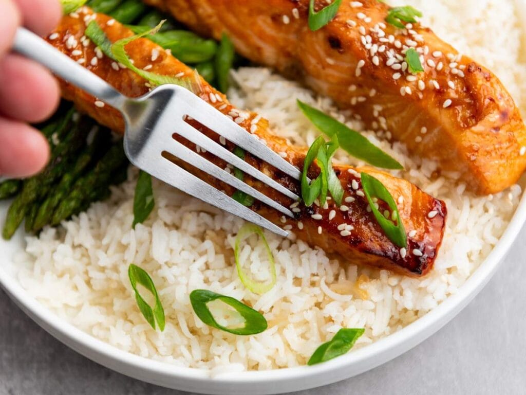 A fork holding a piece of glazed salmon over a plate of white rice, asparagus, and sliced green onions, garnished with sesame seeds.