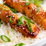 Two glazed salmon fillets topped with sesame seeds and sliced green onions, served over white rice with green vegetables in the background.