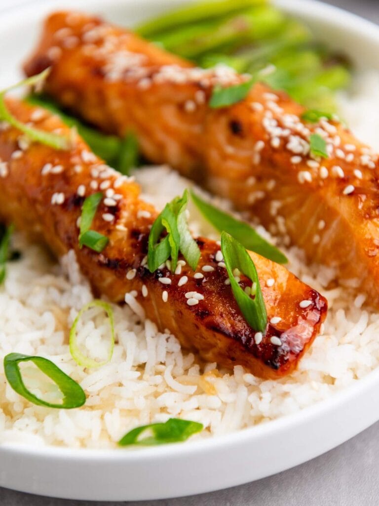 Two glazed salmon fillets topped with sesame seeds and sliced green onions, served over white rice with green vegetables in the background.