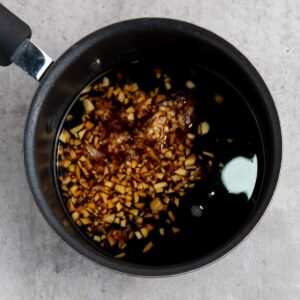 A saucepan containing chopped garlic, soy sauce, and other liquid ingredients on a gray countertop.