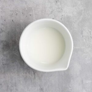 A small white ceramic bowl filled with milk, placed on a light gray textured surface.