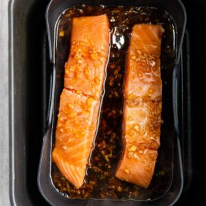 Two raw salmon fillets marinating in a dark sauce with visible pieces of minced garlic in a black tray.