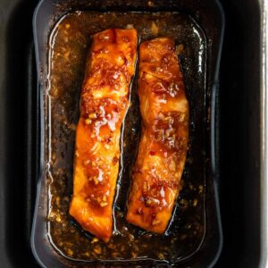 Two glazed salmon fillets with a shiny sauce are baked in a rectangular black tray, surrounded by marinade.