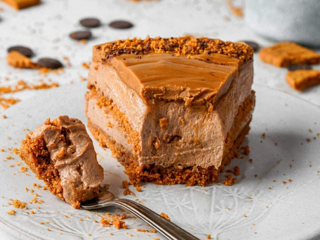 A close-up of a slice of layered chocolate mousse cake with a biscuit crust, topped with caramel glaze, with a fork holding a bite-sized piece in front.