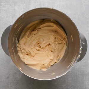 A stainless steel mixing bowl filled with smooth, creamy, light brown frosting on a gray countertop.