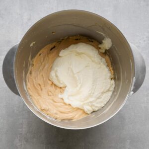 A metal mixing bowl with a light brown batter and a mound of whipped cream being combined, set on a gray surface.