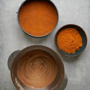 Three containers on a gray surface: one with chocolate batter, one with crushed graham crackers, and one with a graham cracker crust in a springform pan.