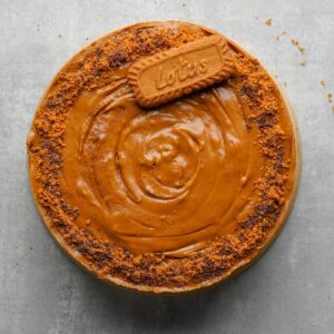 A round cake with caramel-colored frosting, topped with a whole Lotus Biscoff biscuit and crushed biscuit crumbs along the edge, on a gray surface.