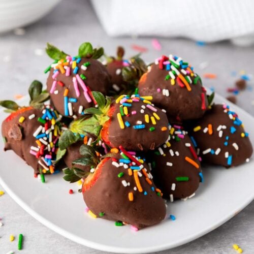 A plate of chocolate-covered strawberries topped with colorful sprinkles, with some sprinkles scattered on the table.