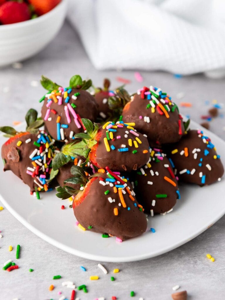 A plate of chocolate-covered strawberries topped with colorful sprinkles, with some sprinkles scattered on the table.