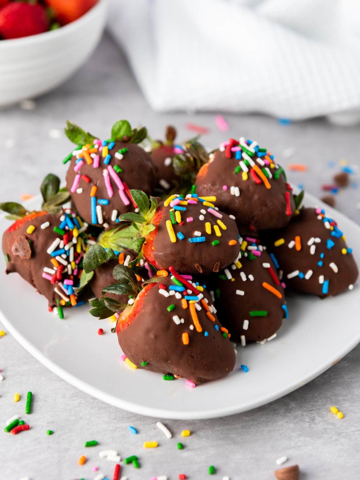 A plate of chocolate-covered strawberries topped with colorful sprinkles, with some sprinkles scattered on the table.