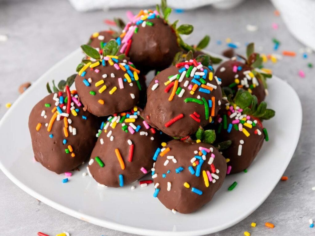 A plate of chocolate-covered strawberries topped with colorful sprinkles, arranged on a white dish.