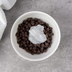 A white bowl filled with chocolate chips topped with a dollop of solid coconut oil, placed on a gray countertop.