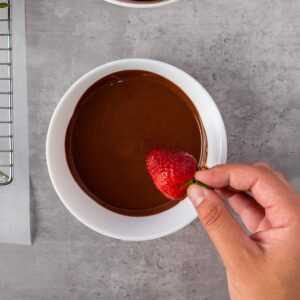 A hand holding a strawberry above a white bowl filled with melted chocolate on a gray surface.