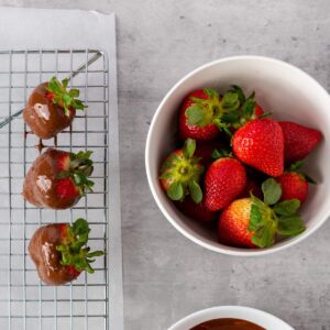 A bowl of fresh strawberries next to a cooling rack with strawberries dipped in chocolate on a gray countertop.