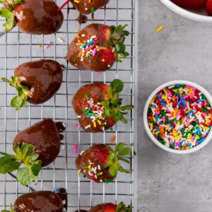 Chocolate-covered strawberries with colorful sprinkles rest on a cooling rack next to a small bowl of rainbow sprinkles on a gray surface.